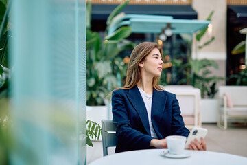 Pensive young businesswoman sitting at a stylish outdoor cafe, holding a smartphone and enjoying a...