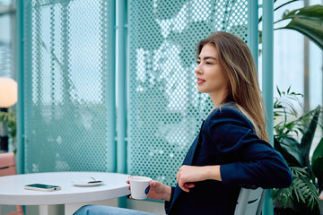 Young woman relaxing and contemplating, sitting at a table in a modern cafe, drinking coffee and looking away