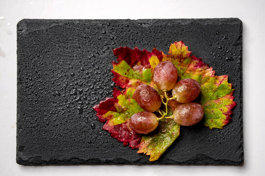 Uvas rojas frescas dispuestas en una superficie de pizarra negra, creando un castraste visual con gotas de agua en las hojas de la parra a&ntilde;aden un aspecto fresco y apetitoso
