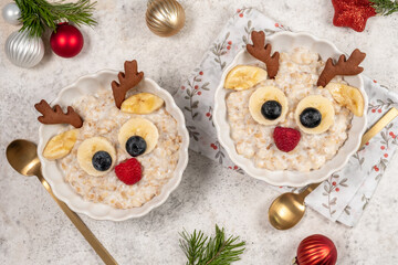 Two bowls of oatmeal designed to look like reindeer are served on a festive table. The bowls are decorated with banana, blueberry, and gingerbread cookie details for a fun breakfast.