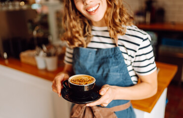 A close-up of a young female barista in an apron holding a freshly brewed coffee in a cozy coffee shop. The hot drink is served in a dark cup. The concept is cozy and atmospheric. Small business.