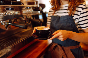 A close-up of a young female barista in an apron holding a freshly brewed coffee in a cozy coffee shop. The hot drink is served in a dark cup. The concept is cozy and atmospheric. Small business.