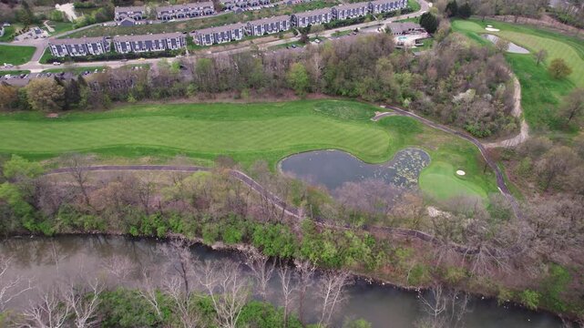 Drone shot of golf course single hole by townhouses, apartments, creek, forest, and green lawn along river by Central Ohio landscape and golfing outing country