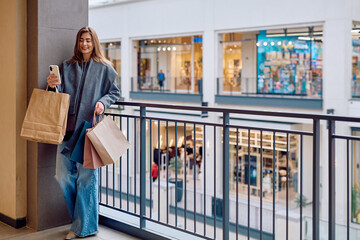 Happy young woman standing on a balcony in a shopping mall, holding several shopping bags and her smartphone