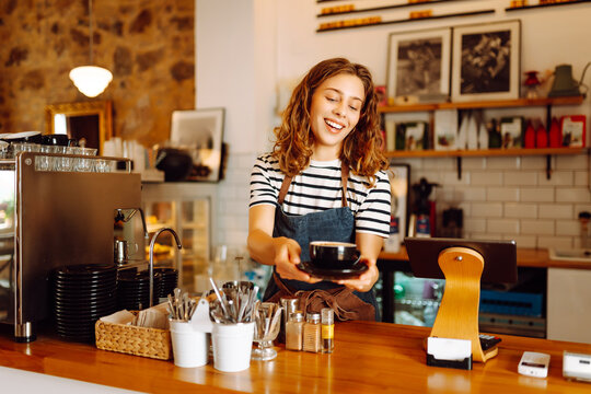 A female barista stands behind the counter with freshly brewed coffee in a cozy cafe. A smiling woman prepares and serves hot drinks inside. The concept is cozy, drinks, and small business.