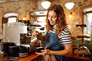 A female barista stands behind the counter with freshly brewed coffee in a cozy cafe. A smiling...