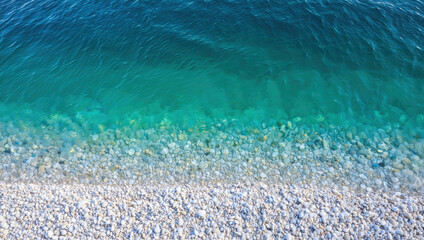 Clear turquoise water meets a smooth pebbled beach on a sunny day. The gentle waves create a soothing sound while the sunlight reflects off the water surface, enhancing the serene atmosphere