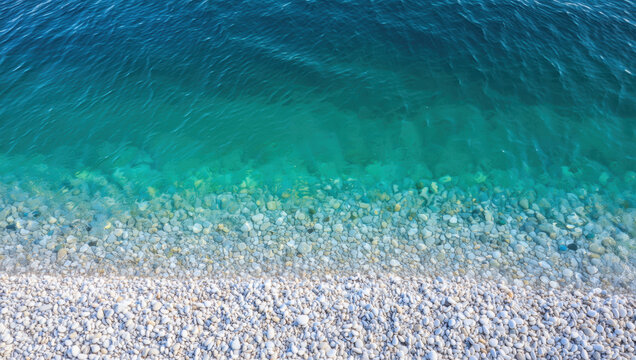 Clear turquoise water meets a smooth pebbled beach on a sunny day. The gentle waves create a soothing sound while the sunlight reflects off the water surface, enhancing the serene atmosphere
