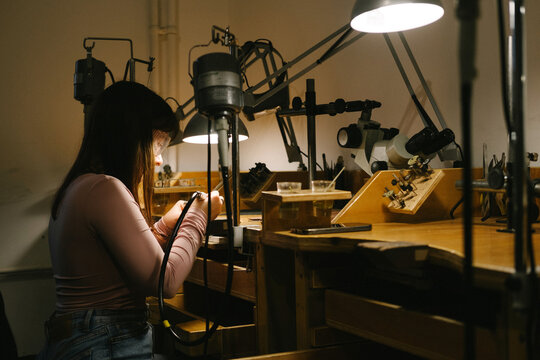 woman jeweler workshop workbench tools lamp jewelry craftsmanship focused on making a handmade ring at a wooden bench with soldering iron task lights and warm light