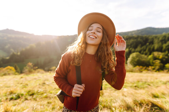 A happy woman wearing a hat and carrying a backpack enjoys the green hills at sunset. A traveler strolls and admires the mountain landscape. Concept of adventure and relaxation.