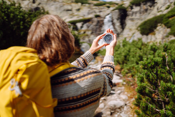 Rear view of female hiker, holding backpack and compass, on rocky trail in background. Hiker looks for destination and enjoys the mountain scenery on a sunny day. Concepts of nature, navigation.