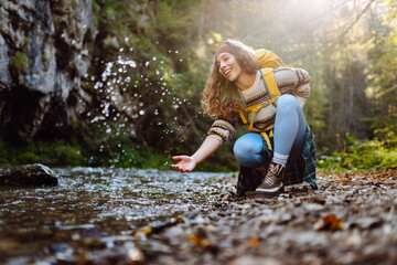 A female hiker with yellow backpack touches water near mountain river surrounded by forest. Beautiful woman on hiking trail in  forest on sunny day. Concepts of hiking, adventure,  nature.