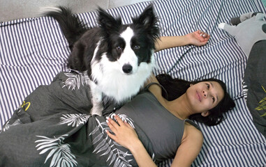 A close-up scene shows a Border Collie dog sitting upright on a bed, with a young woman lying beneath it and looking at the camera, capturing a moment full of playfulness and connection.