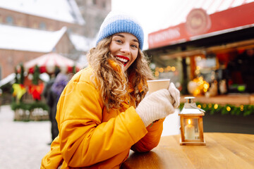 A cheerful woman in a bright jacket holds a hot drink at an atmospheric winter market. A young...