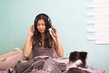 With closed eyes and headphones, the woman sits peacefully on the bed, enjoying a moment of musical solitude while her Border Collie sits nearby, both appearing serene.