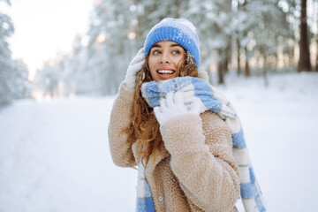 Obraz premium A curly-haired woman in warm clothing is having fun in a snowy forest at sunset. A young woman enjoys a sunny day outdoors. Nature concept, walk. Winter day.