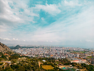 A serene aerial panorama of a large coastal city framed by soft blue skies, distant sea, and surrounding mountains. Natural daylight and calm atmospher