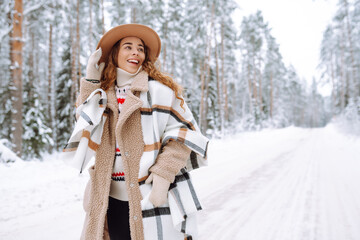 Fototapeta premium A cheerful woman stands on snowy path, wearing warm clothing. A hiker in a hat enjoys a snowy day outdoors in the forest. Nature, hiking, and lifestyle concepts.