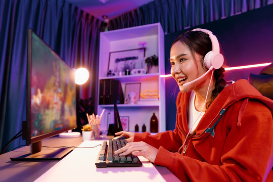 Host channel of young beautiful Asian gaming streamer playing fighting Moba at battle arena game on pc screen with multiplays, wearing pastel headphone at neon digital light modern room. Stratagem.