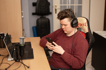 Young man sitting at a cluttered desk with headphones on, distracted from his laptop and microphone as he browses his smartphone instead of working.