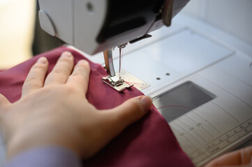 A seamstress's hands work on burgundy fabric on a sewing machine in a studio.