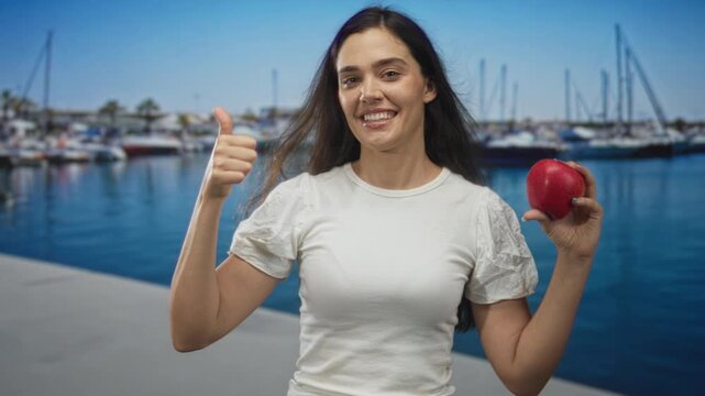 Woman holds red apple and points finger to it at a busy port dock with moored boats; healthy eating happiness.