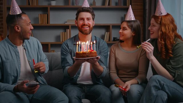 Four multiracial cheerful friends at home party congratulate birthday man laughing carefree smiling holding cake with candles happy diverse group men women surprise event celebration festive meeting 