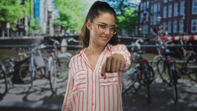 Young hispanic woman fist to chest brotherhood gesture on street in amsterdam canal area with parked bicycles and water; solidarity support.