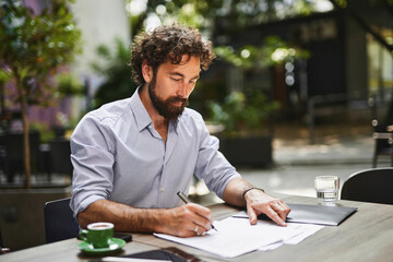 A man with curly hair and a beard sits at a table in a contemporary cafe, focused on writing notes and examining paperwork while enjoying a cup of coffee.