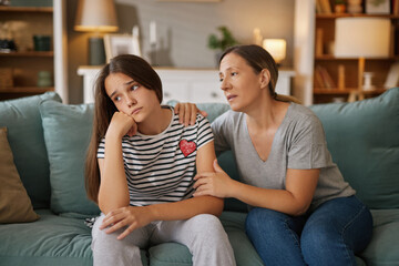 A mother offers support to her teenage daughter who looks troubled. They sit together on a couch in a warm living room filled with soft lighting, fostering a comforting atmosphere.