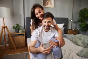 A couple shares a moment of laughter and connection as they gaze at a smartphone in their comfortable, well-decorated bedroom during a relaxed afternoon.