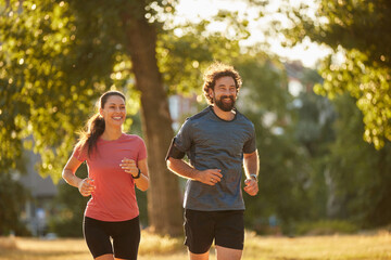 Two joggers run together in a sunny park, smiling and enjoying their workout. The scene captures their commitment to health and active lifestyle amid nature.
