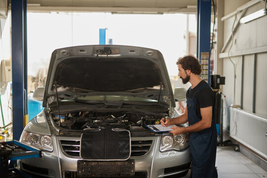 A car mechanic examines the engine of a silver vehicle while holding a clipboard in an auto repair garage. The shop is well-lit and organized.