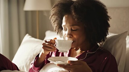 happy african american woman drinking tea in bed. lady wearing silk pajamas holding cup with hot beverage. morning sunlight and relaxation concept, lifestyle