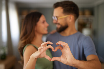 A loving couple creates a hand heart gesture while embracing at home, showcasing their affection and connection in a cozy indoor setting.