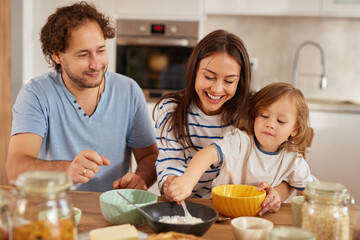 In a warm kitchen, a family shares joy as they cook together. Parents smile at their child, who playfully helps mix ingredients. The atmosphere is filled with love and fun.