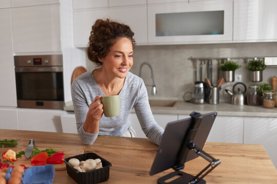 A woman sits at a wooden kitchen counter, smiling while holding a mug of coffee. She watches a cooking video on a tablet, surrounded by fresh ingredients like vegetables and eggs.
