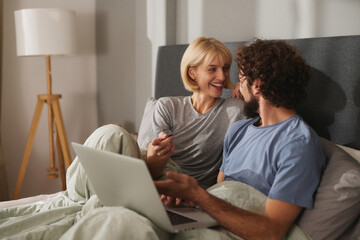 A man and woman cuddle under blankets in bed, smiling and chatting while using a laptop, enjoying each other's company during a quiet evening at home.