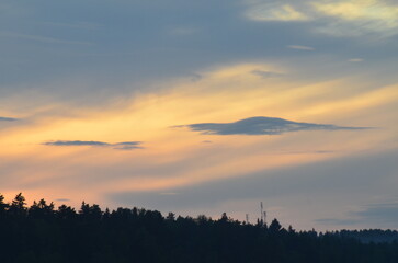 Obraz premium The sun behind clouds. A sunny evening, a sky with small clouds through which the bright sun shines. Beneath the clouds, a coniferous forest grows like a dark wall, the tops of tall trees are visible.