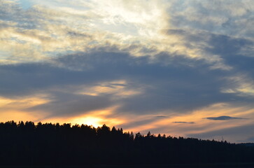 The sun behind clouds. A sunny evening, a sky with small clouds through which the bright sun shines. Beneath the clouds, a coniferous forest grows like a dark wall, the tops of tall trees are visible.