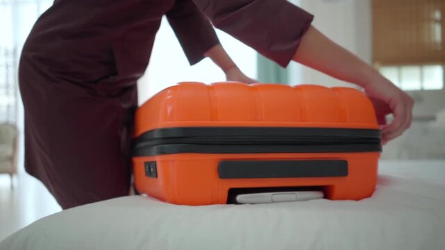 A woman closes a bright orange suitcase on a bed, showing a close view of her hands and the travel case in a clean home interior.