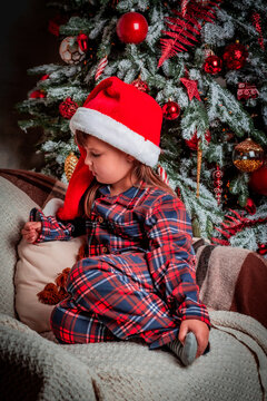 Happy little girl child in pajamas and festive santa hat smiling while sitting near christmas tree. Magic. New Year's eve. St. Nicholas Day. Present. Time of holidays and miracles.	