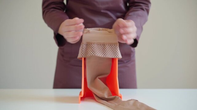 A woman stretches a compression stocking over an assist device on a table, showing a close view of her hands and the fabric prepared for medical leg support.