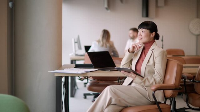 A professional Asian woman in smart attire engages with her laptop in a modern workspace while her colleagues collaborate nearby.