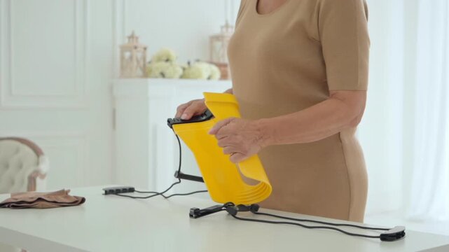 A senior woman holds a yellow assist device on a table, preparing it for use with compression stockings in a bright and clean home setting.