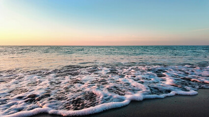 Gentle ocean waves softly approach sandy beach, creating calming seaside scenery during colorful sunset sky.