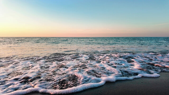 Gentle ocean waves softly approach sandy beach, creating calming seaside scenery during colorful sunset sky.