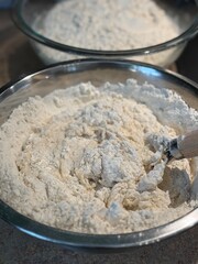 Two mixing bowls full of the ingredients to make loaves of sourdough bread. The dough is in the process of being mixed and lots of lose flour is still visible in the bowls. 