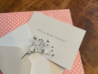 Fototapeta premium A white paper card next to its matching white envelope with the printed text “Will you be my Valentine?” with a pink gift box below. A sprig of fresh baby’s breath flowers rests on top. 