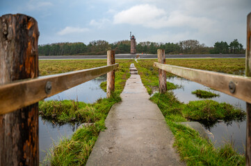 A concrete pathway leads through a grassy area with a wooden fence, leading to distant Boehler Lighthouse, surrounded by trees under a cloudy sky. St Peter Ording, North Sea, Germany